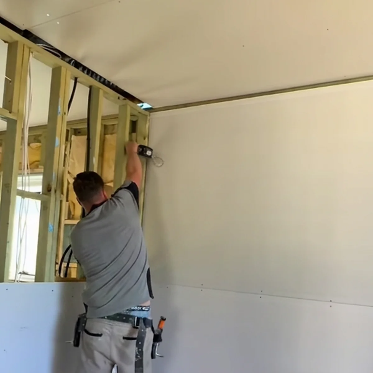 A plasterer screwing off a wall sheet in a room being lined, with the ceiling already sheeted and the timber wall studs still open alongside.