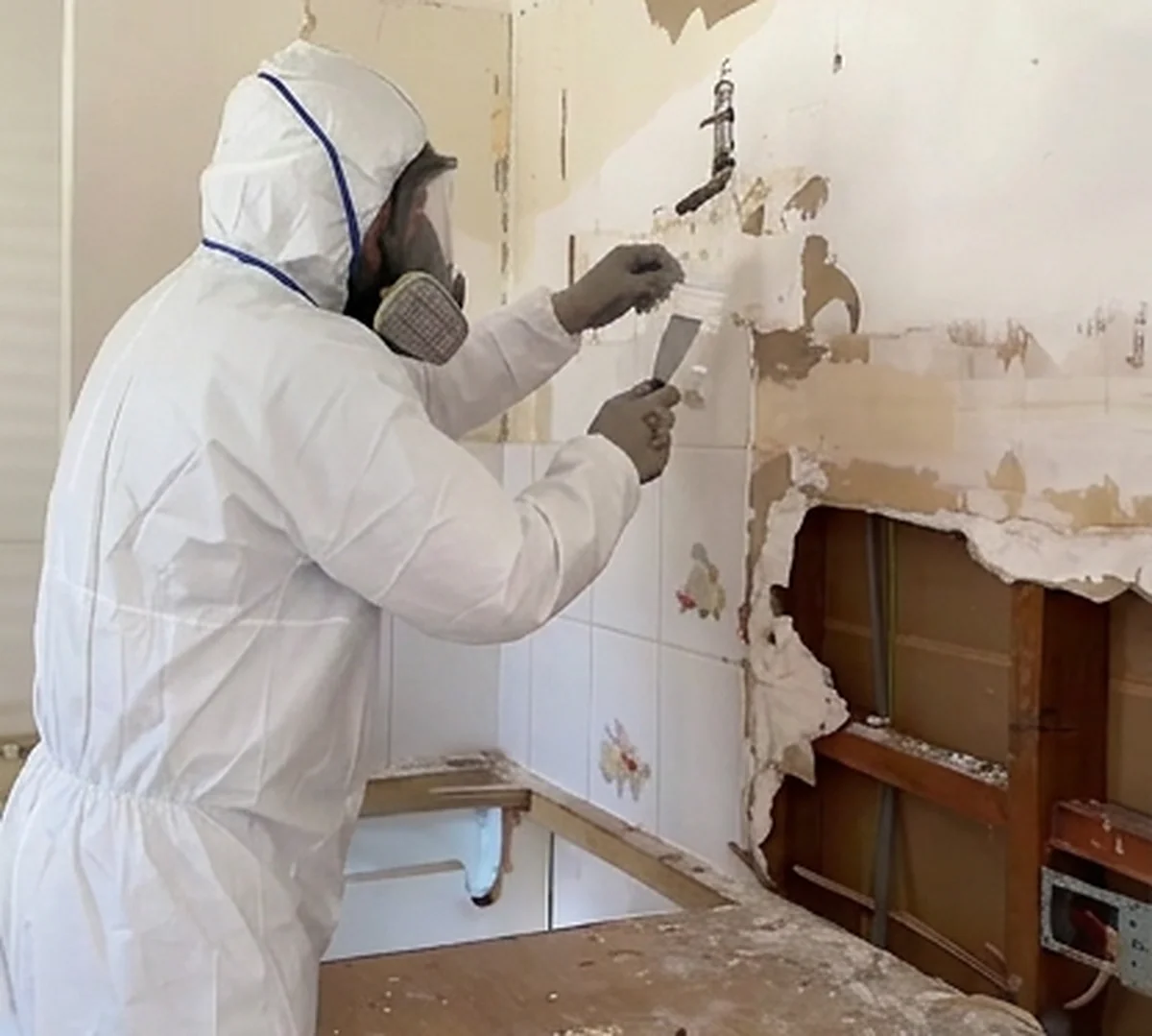 A person in a full protective suit and respirator stripping tiles from a bathroom wall, with the framing behind already exposed.