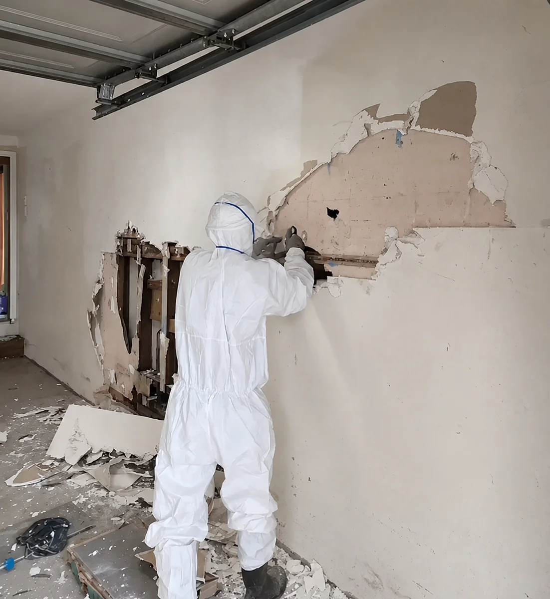 A person in a full protective suit pulling lining from a wall below a roller door track, with broken sheeting across the floor.
