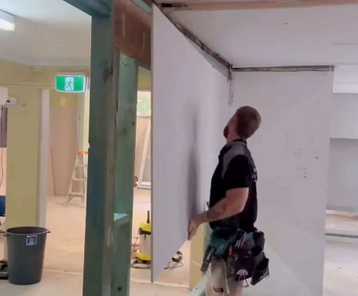 A plasterer fixing a sheet to a wall in a shop fitout, with an exit sign and stacked sheets behind.