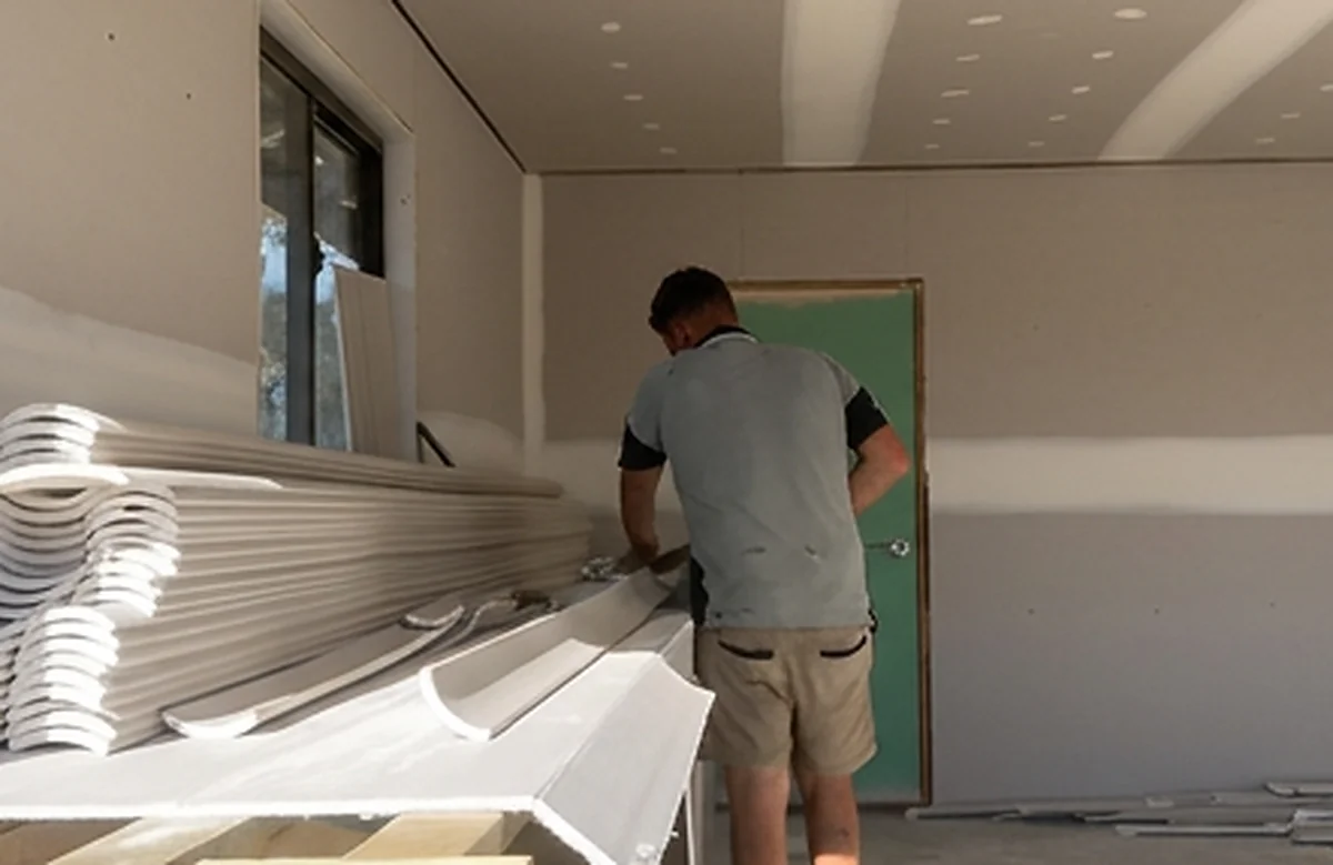 A plasterer working at a stack of cornice lengths on trestles in a newly sheeted room.