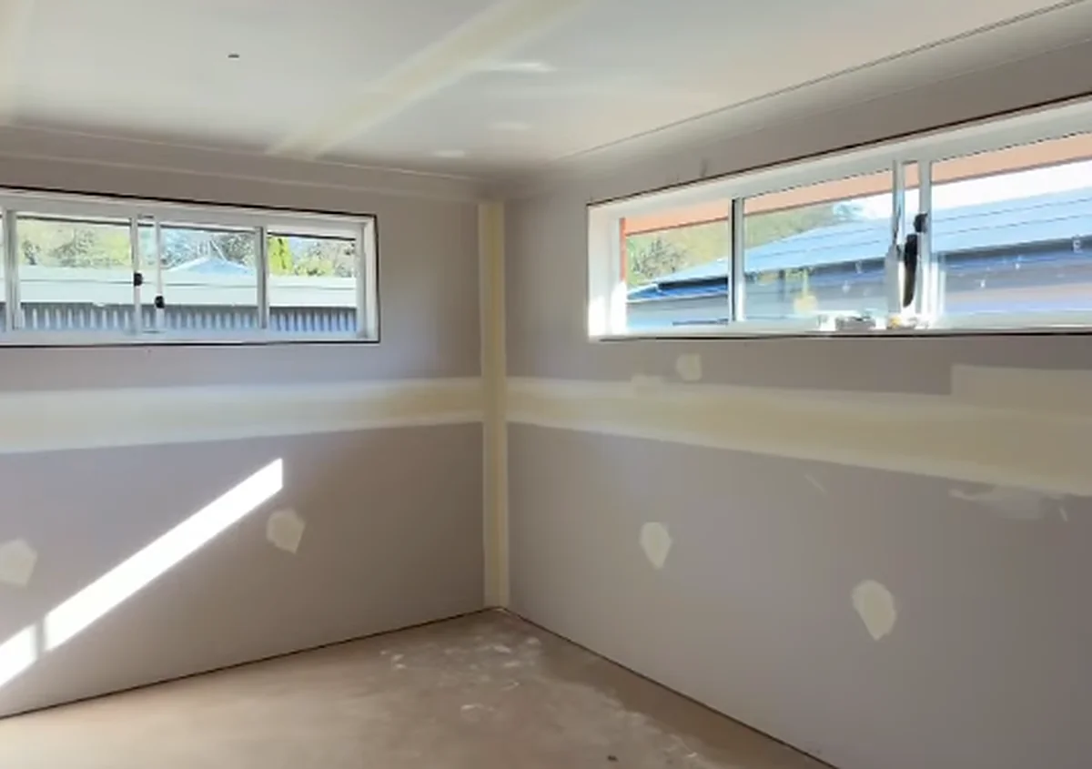A sheeted bedroom in a new house, walls and ceiling jointed and spotted, with sunlight falling across the bare concrete floor.