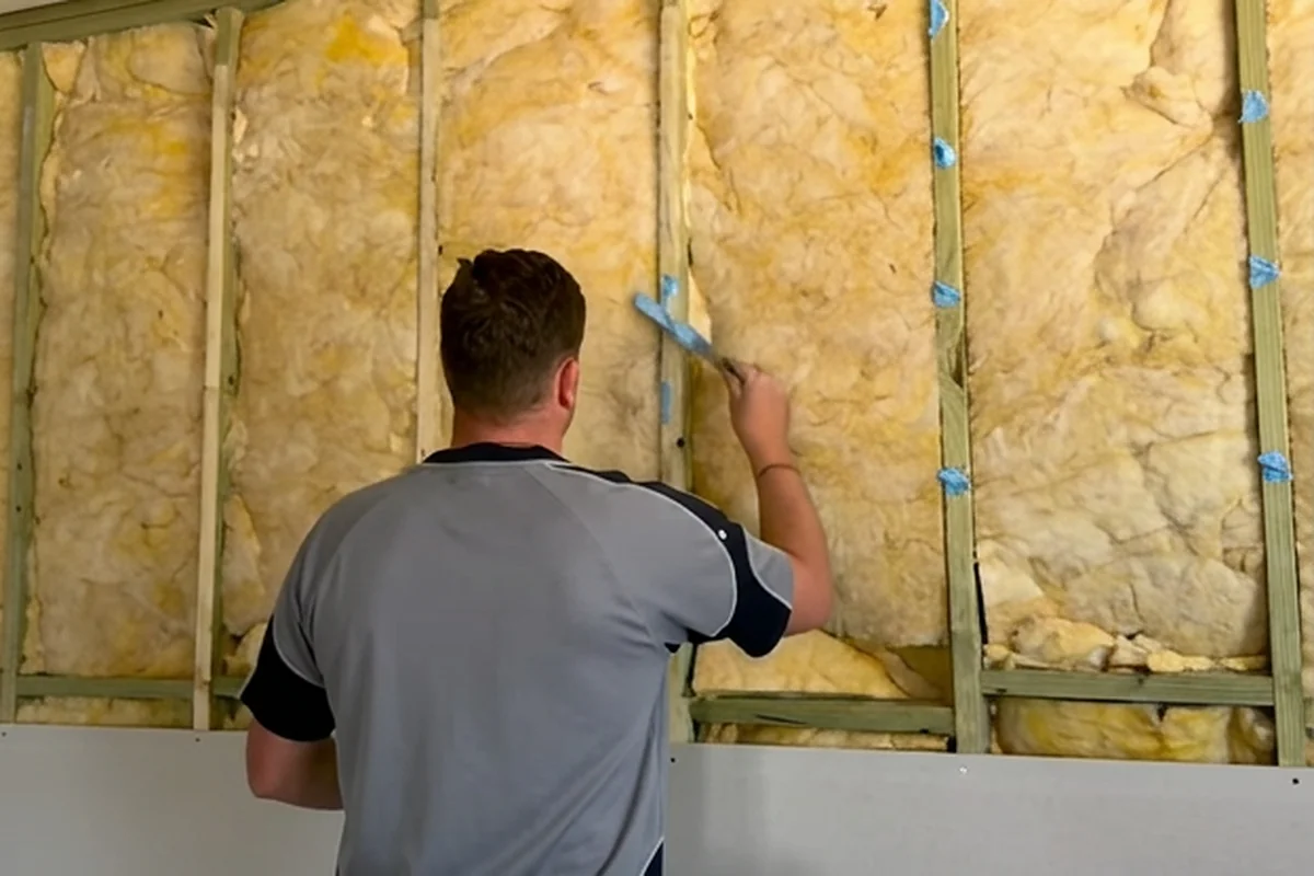 A plasterer at an open timber stud wall packed with insulation batts, with the first row of sheets already fixed along the bottom.