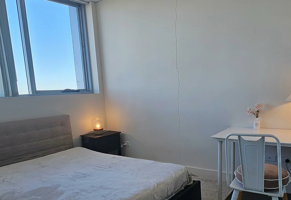 A crack running down a bedroom wall from the ceiling towards the skirting, beside the bed and the window.