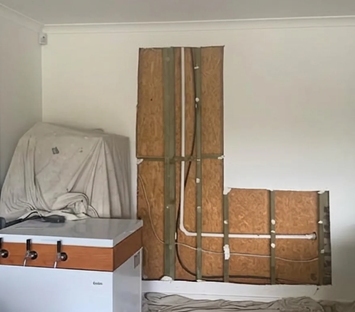 A wall opened up to the framing with the studs and pipework exposed, beside a laundry tub under a drop sheet.