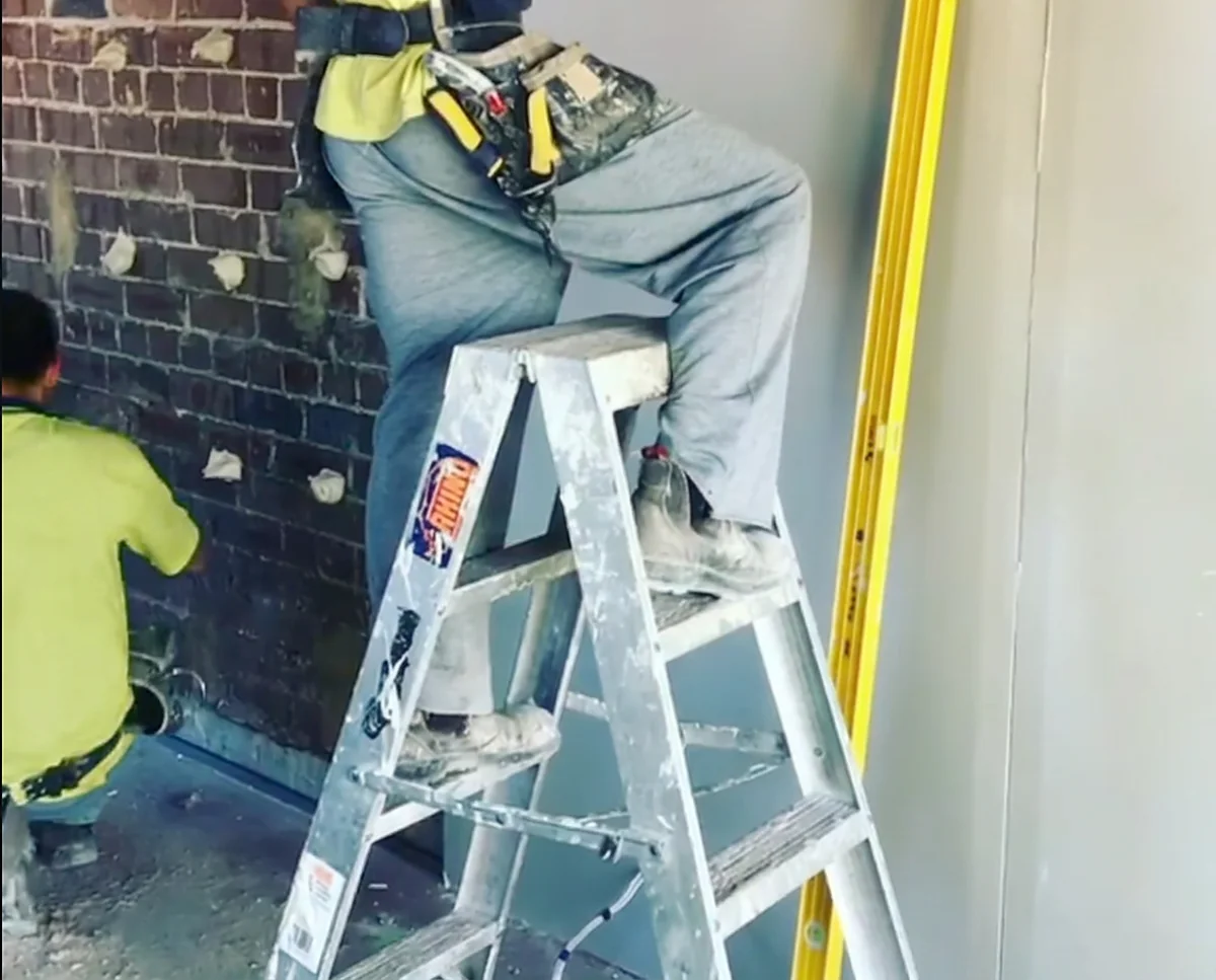 A plasterer on a stepladder pressing a sheet onto an old brick wall, with daubs of adhesive spaced across the brickwork.