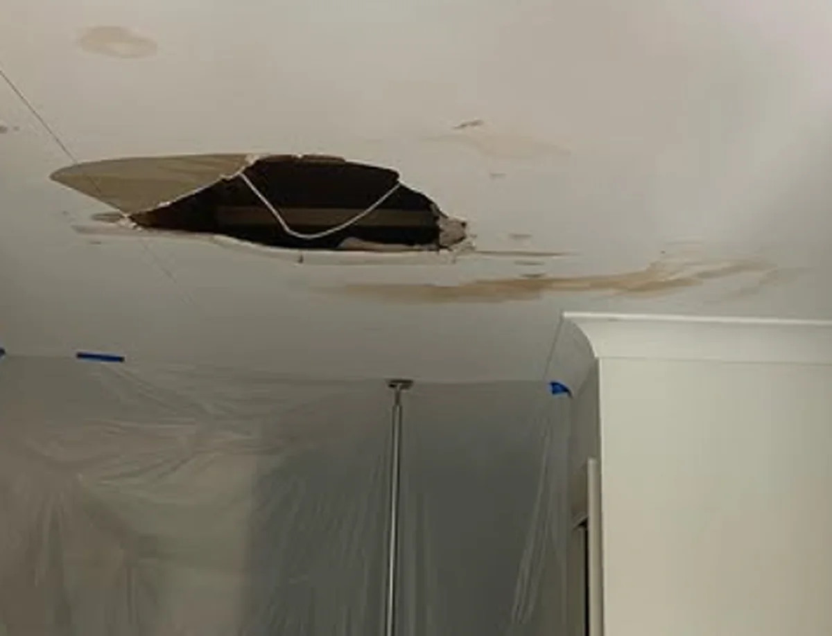 A ceiling with a jagged hole punched through it and brown water staining spreading around it, with plastic sheeting hung below.