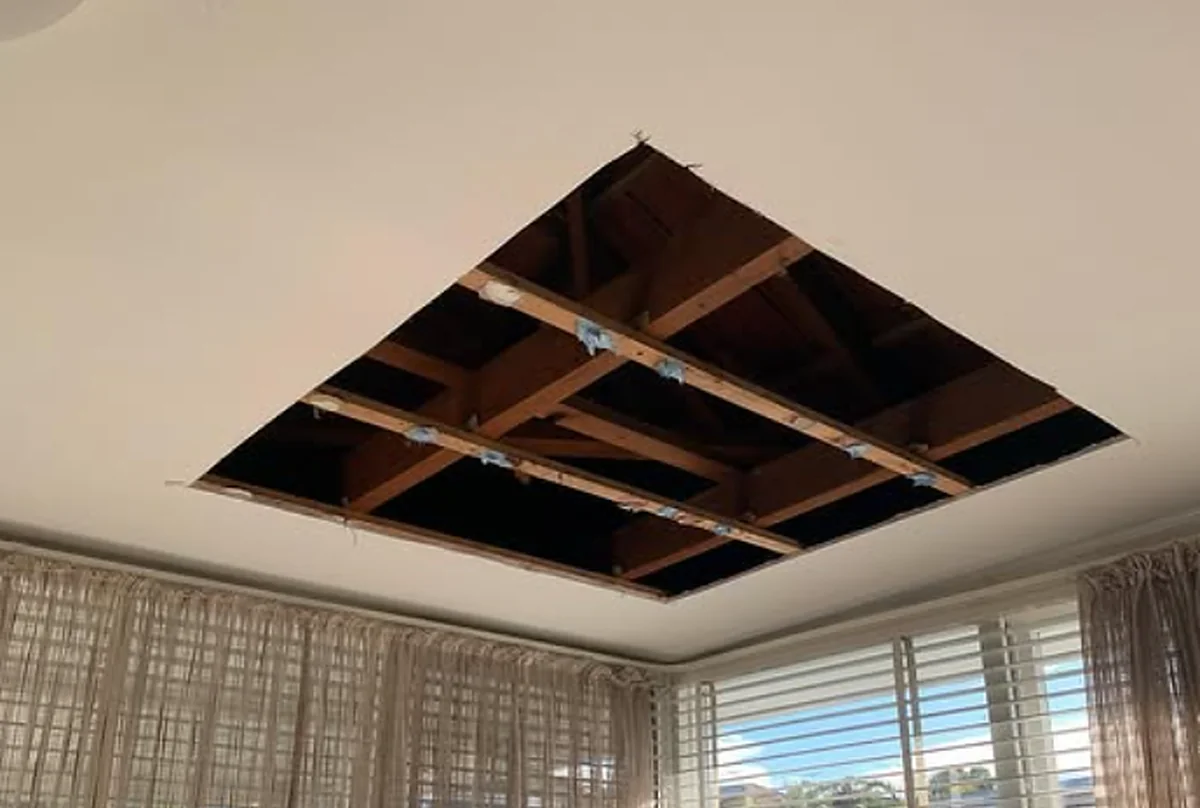 A square opening cut out of a bedroom ceiling, showing the timber joists and battens above with the plaster removed.